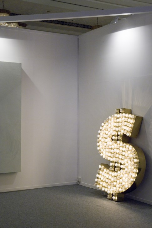 Illuminated dollar sign sculpture made of light bulbs mounted on a gallery wall