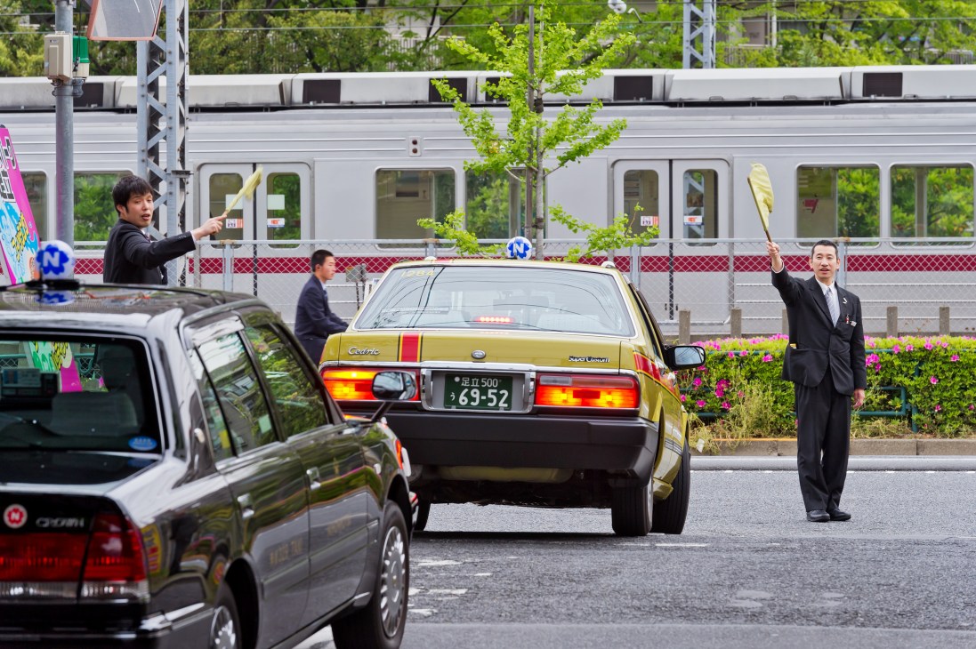 Tokyo taxi drivers in black suits directing traffic as yellow and black taxis depart from depot near train station.