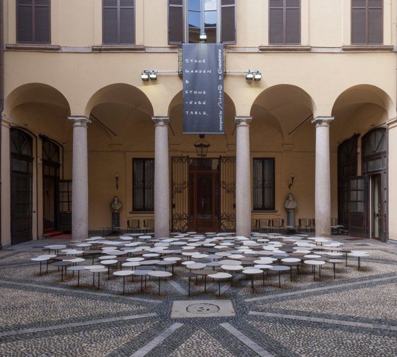 Nendo's one-legged stone tables installed in a Milan palazzo courtyard with arched columns.