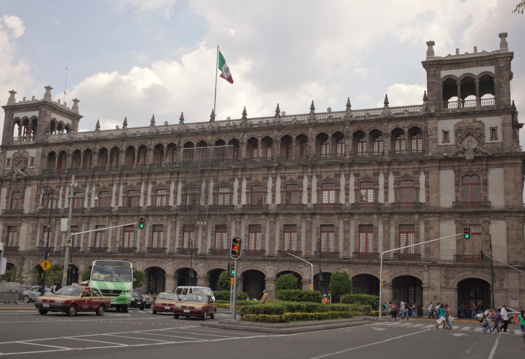 Government Palace in Mexico City with Mexican flag, colonial architecture, and street traffic below.