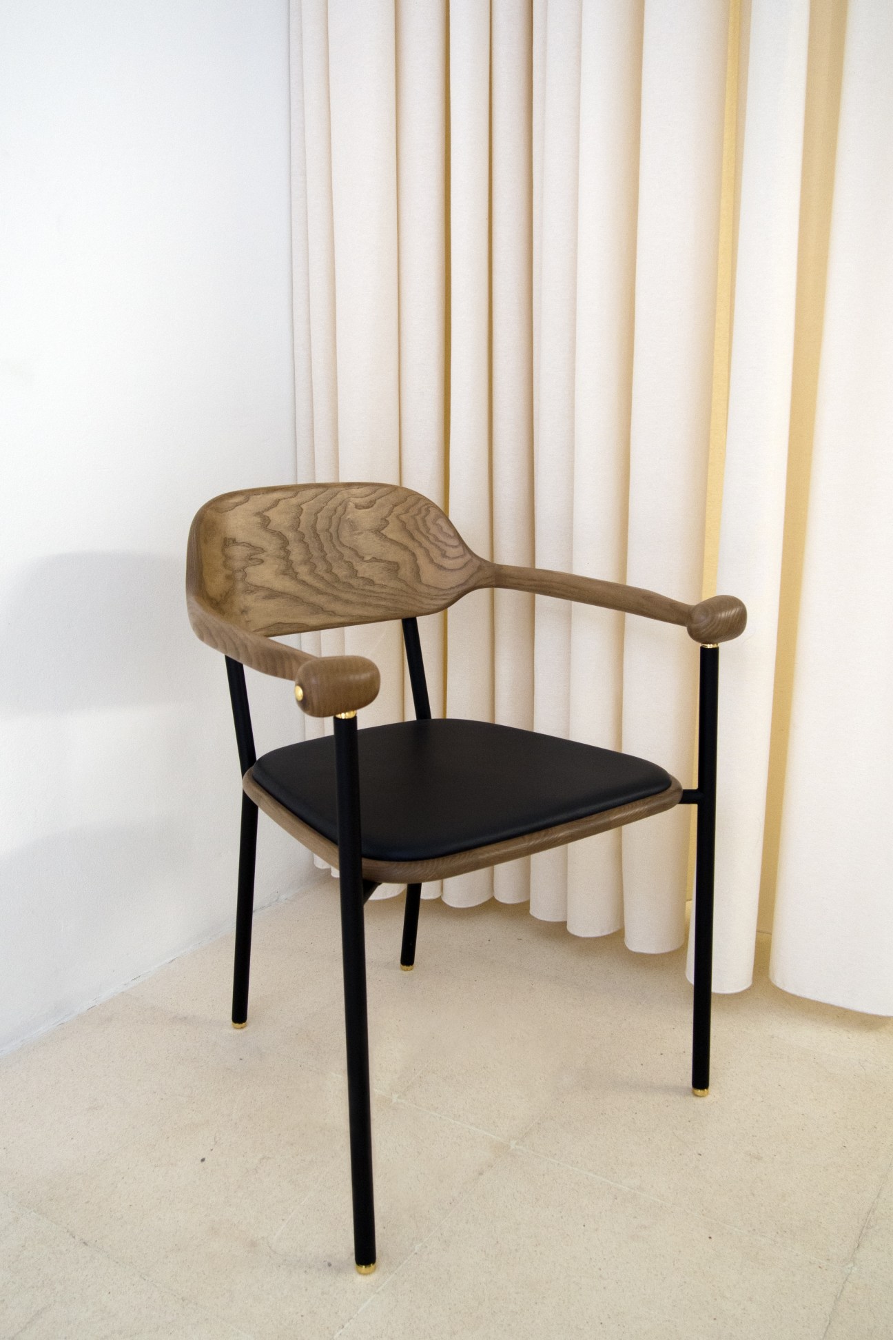 Wooden armchair with carved backrest, black metal legs, and black seat cushion against cream curtains.