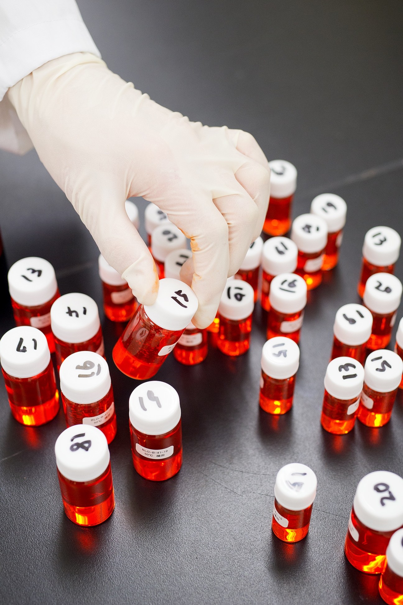 A gloved hand holds a numbered sample vial of red liquid among rows of similar test samples on a dark surface.
