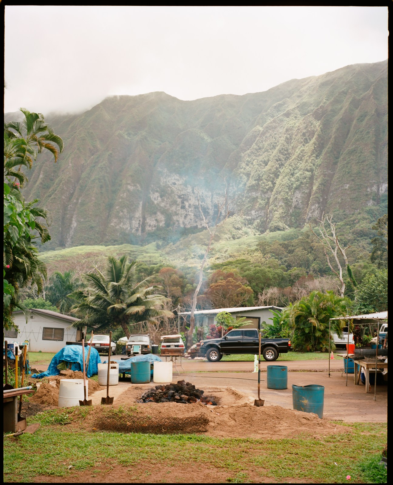 Fire pit surrounded by metal containers in Pu'uhonua O Waimanalo village with mountains and homes in background.