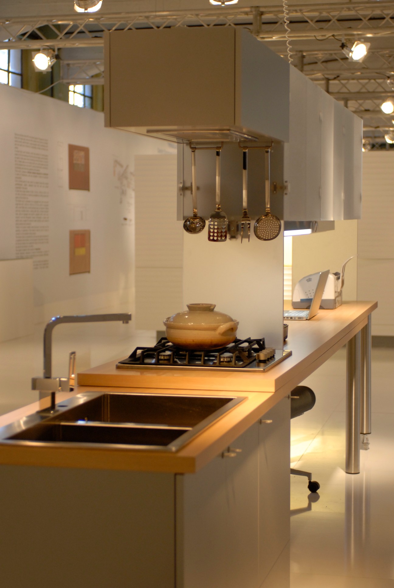 Modern kitchen exhibition with minimalist design, gas cooktop, sink, and hanging cookware display.