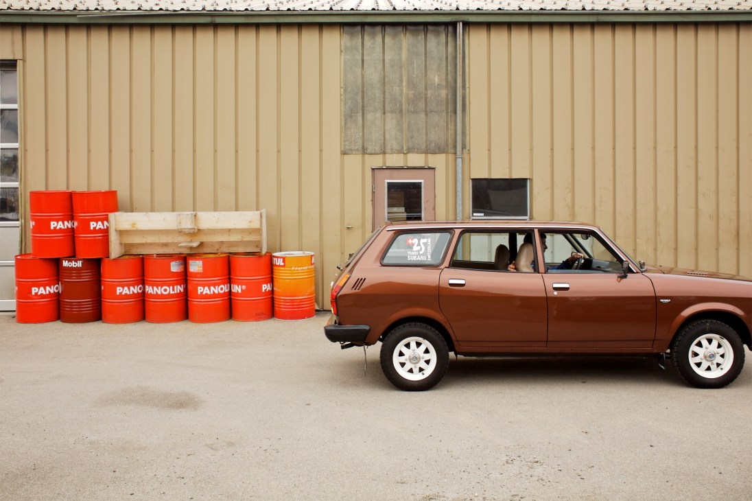 Rita Holmberg-Bertschi in a brown Subaru A67 1600 from 1979 parked beside red oil drums.