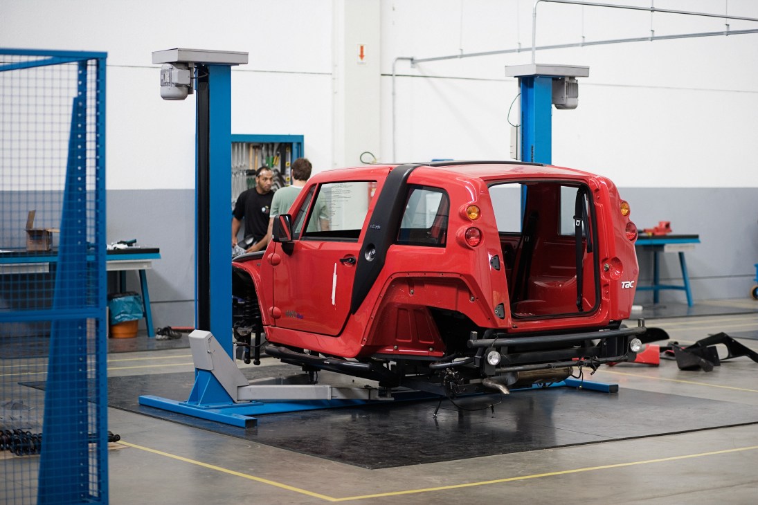 Red vehicle undergoing assembly or inspection in an industrial workshop with blue equipment and metal framework.