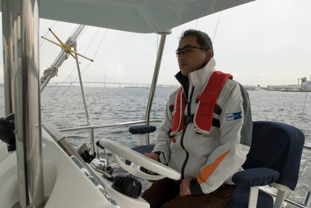 Man at the helm of a sailboat wearing a life jacket, with Yokohama Bay visible behind him.