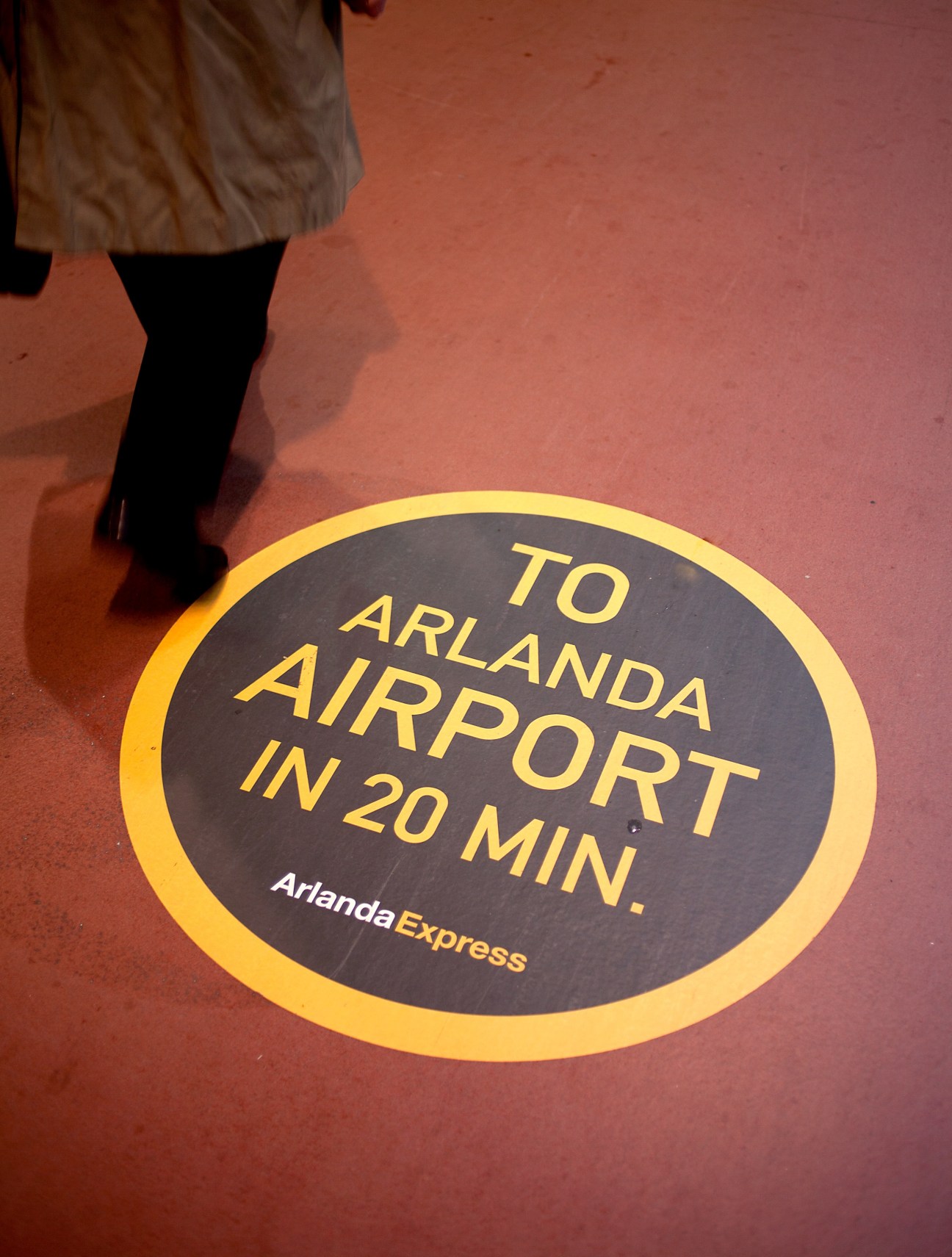 Yellow circular wayfinding sign on red floor directing passengers to Arlanda Airport in 20 minutes.