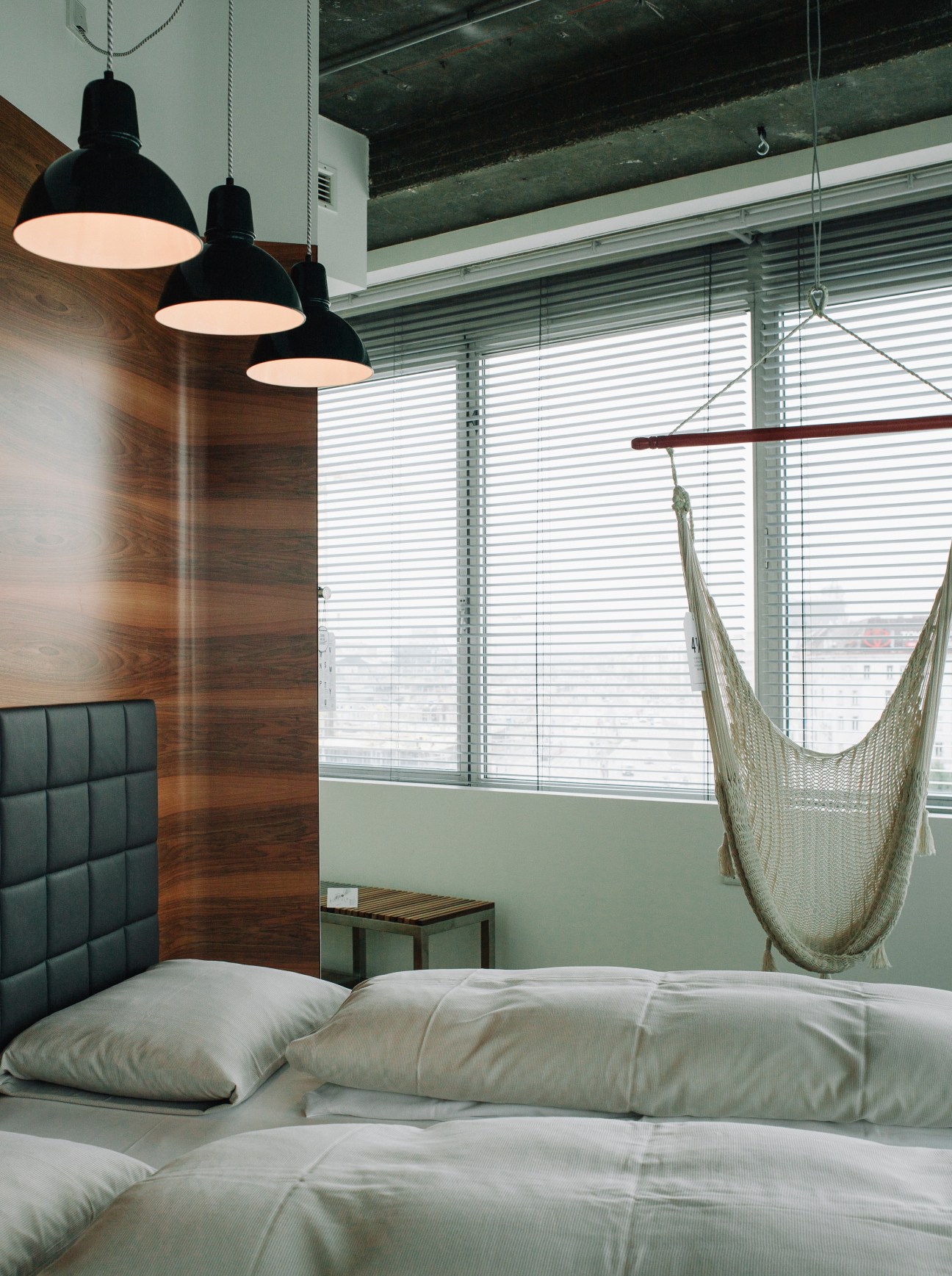 Modern hotel bedroom with wooden accent wall, black pendant lights, hammock chair, and green upholstered headboard.
