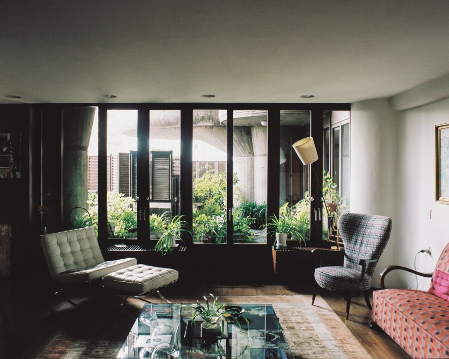 Modernist living room with black-framed windows overlooking a garden courtyard, vintage furniture including patterned chairs, and brass floor lamp.