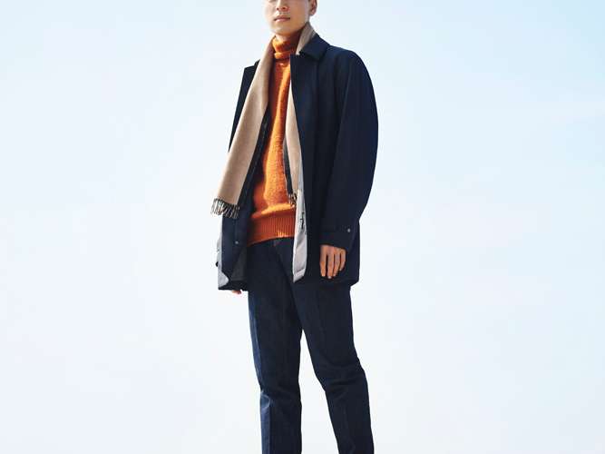 Model wearing layered winter menswear in neutral tones including coat, rollneck jumper and scarf