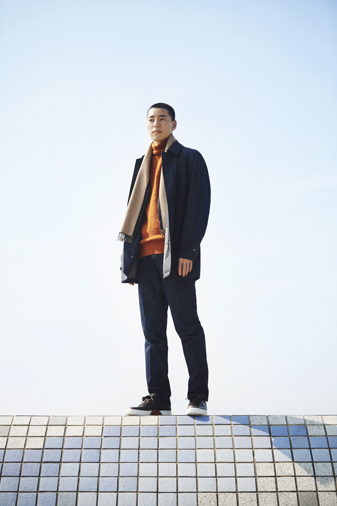 Model wearing layered winter menswear in neutral tones including coat, rollneck jumper and scarf