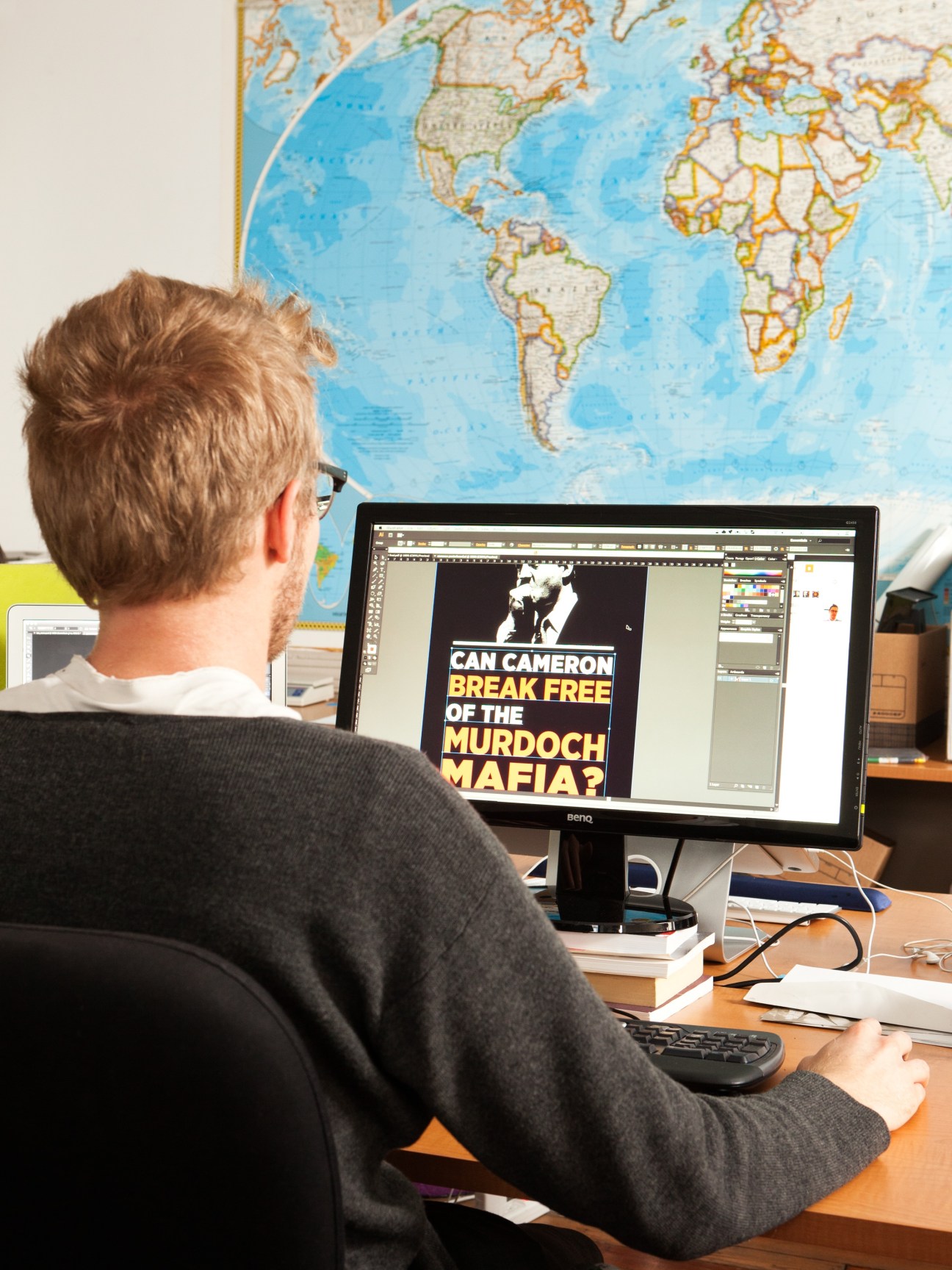 Man at desk working on computer displaying activist campaign graphic about breaking free from Murdoch media influence, with world map on wall behind.