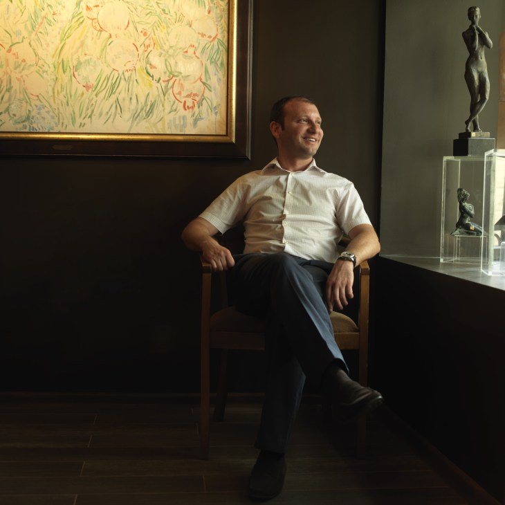 Emin Mammadov, art dealer, sits in a gallery surrounded by artwork including a framed painting and sculptures.