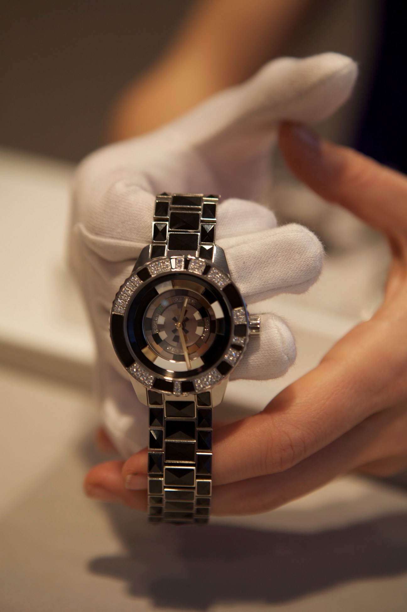 A person in white gloves holds Dior's Christal Mystérieuse watch with black ceramic case and metal bracelet.
