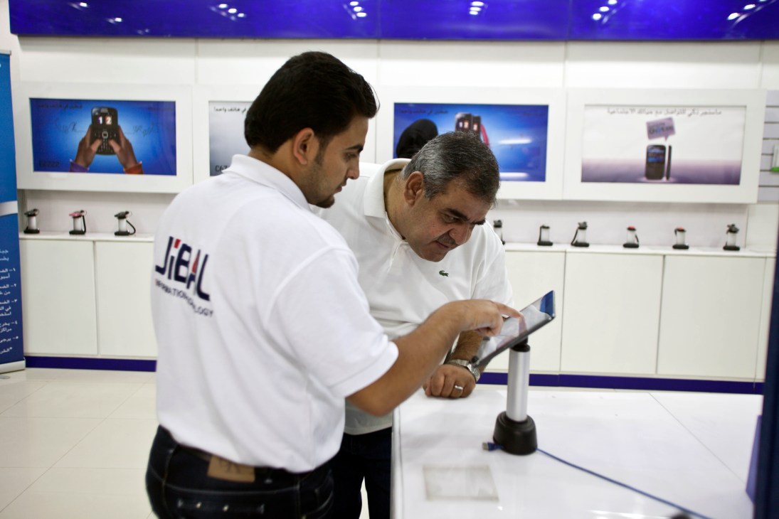 Two men examine a tablet device in an electronics store displaying phones and products.