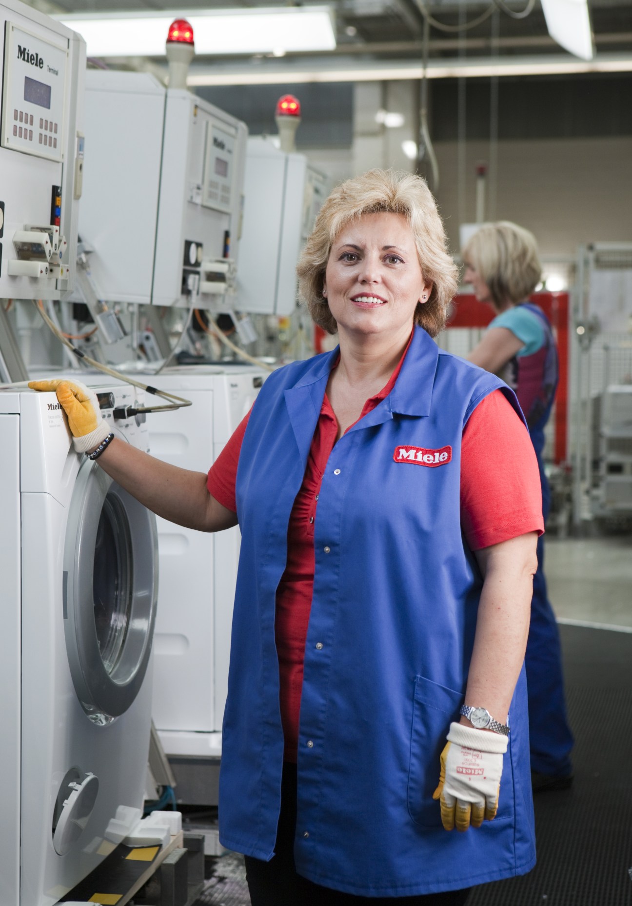 A Miele factory worker in branded blue and red uniform stands at a washing machine workstation.
