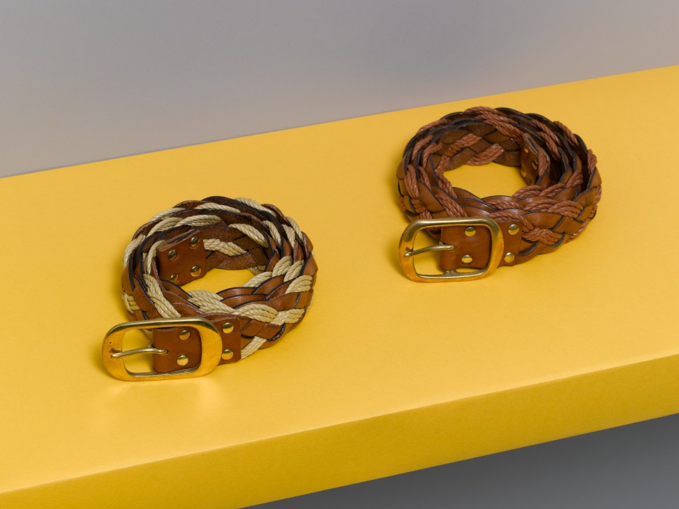 Two woven leather belts with brass buckles displayed on a yellow surface against a gray background.