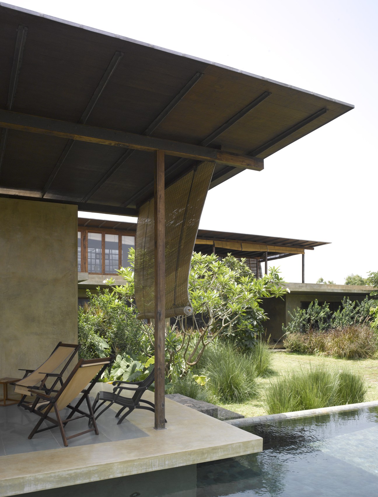Modern covered pavilion with wooden pergola and betel-nut wood screens overlooking a pool and landscaped garden with native plants.