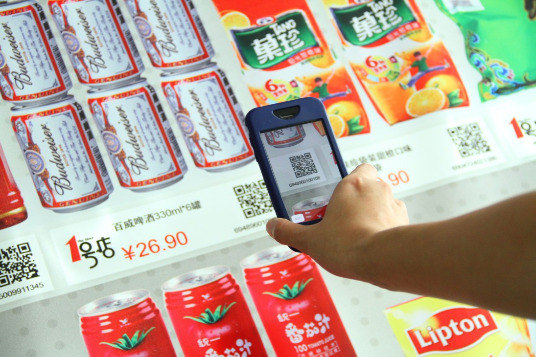 Hand holding smartphone scanning QR code on LED screen displaying product barcodes and prices in Shanghai subway virtual supermarket.