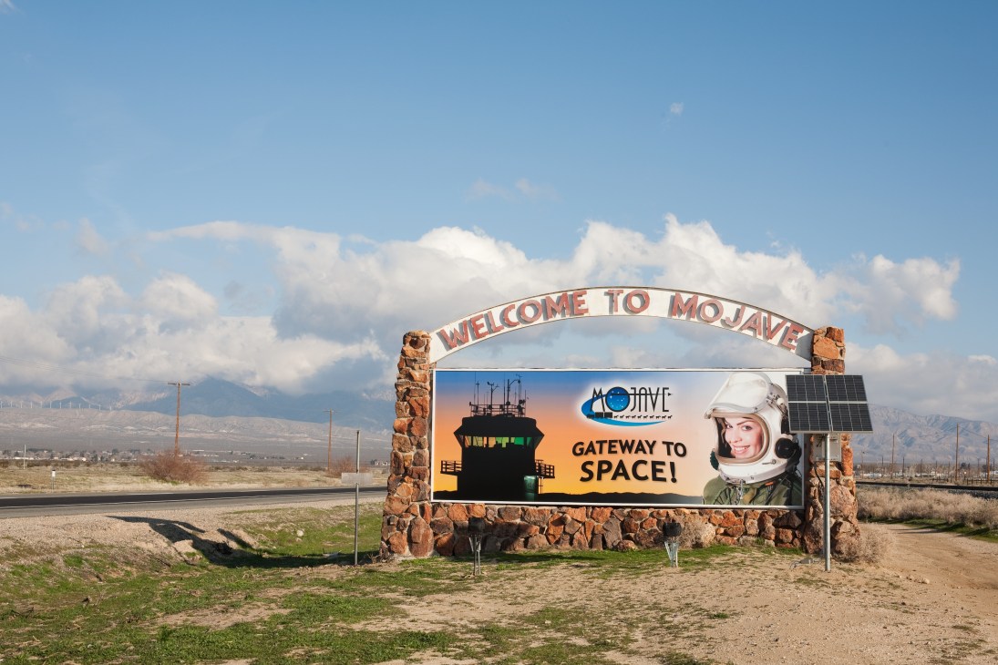Welcome to Mojave Gateway to Space sign with astronaut illustration and solar panel, desert mountains in background.