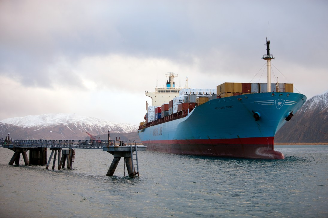 A blue and red cargo ship laden with shipping containers approaches a dock at Dutch Harbor with snow-capped mountains in the background.