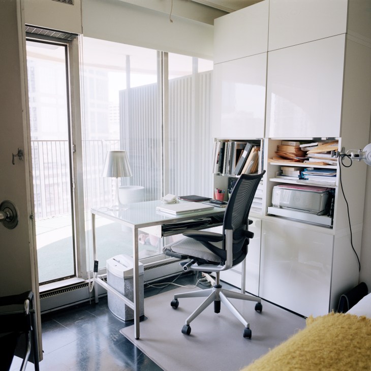 A minimalist bedroom study with white built-in shelving, a desk, ergonomic chair, and floor-to-ceiling window opening onto a balcony.