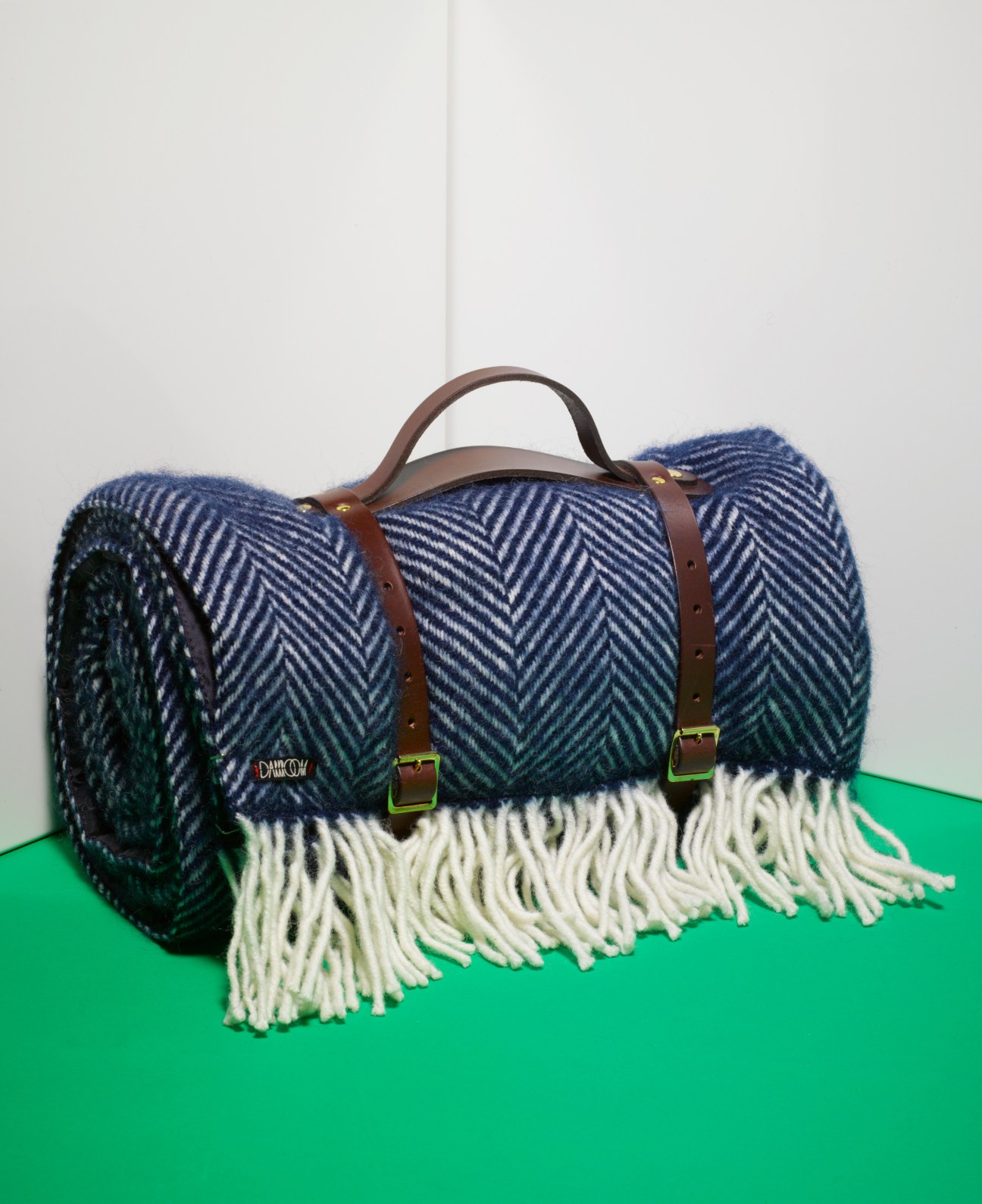 Darkroom tartan picnic blanket rolled with leather handles and cream fringe on green surface.