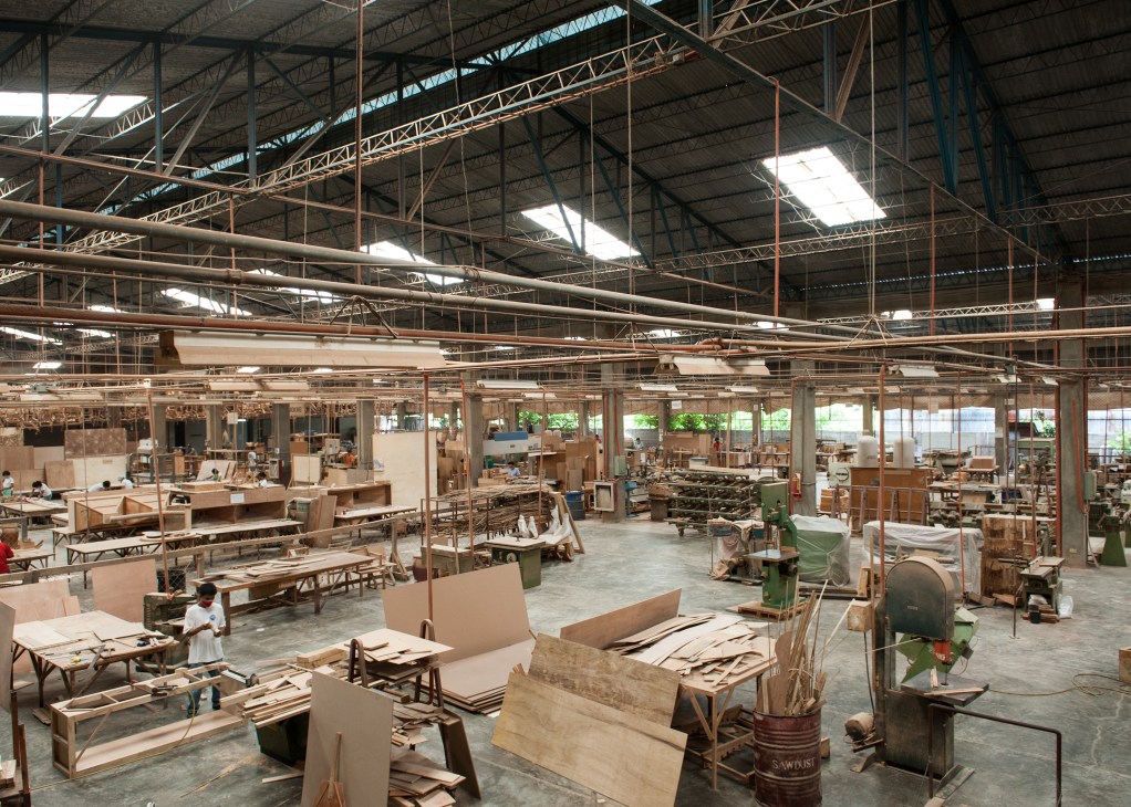 Large industrial furniture factory floor with wooden furniture pieces in production, metal structural beams overhead, and workers throughout the space.