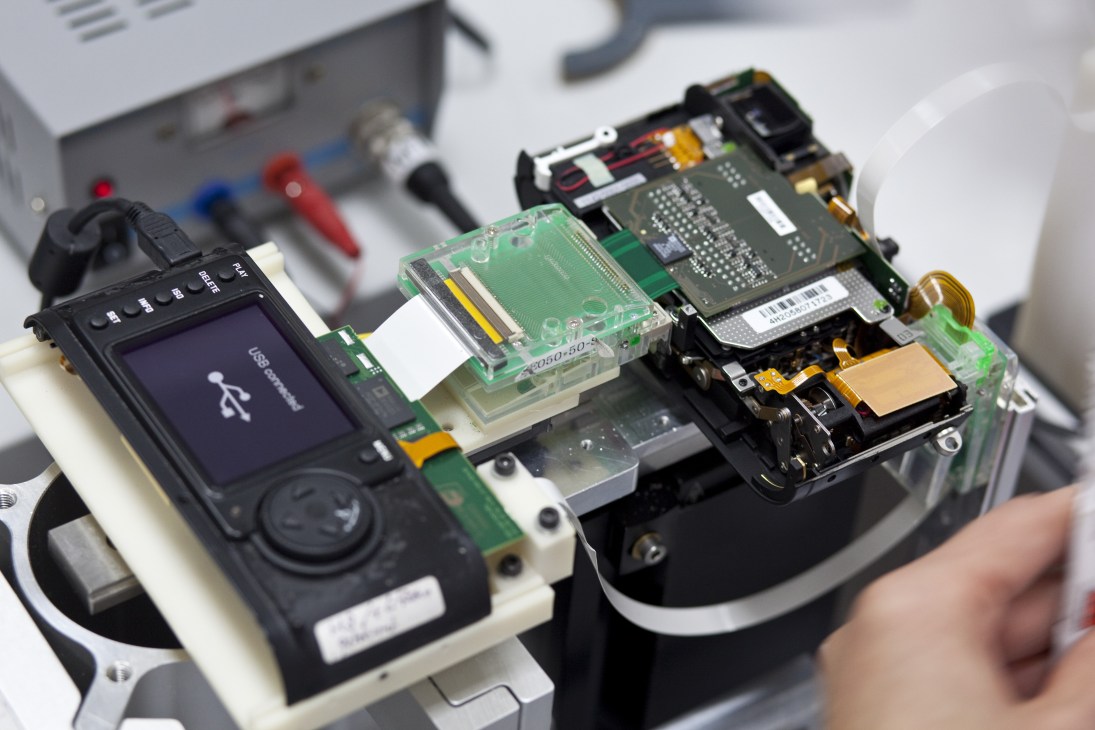Disassembled Leica camera components including circuit boards, lens, and internal mechanisms displayed on white surface.
