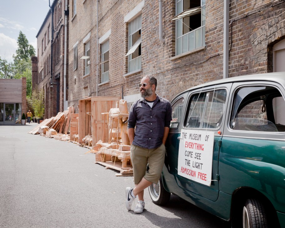 Man standing beside vintage van labeled 'The Museum of Everything' with wooden sculptures stacked nearby.