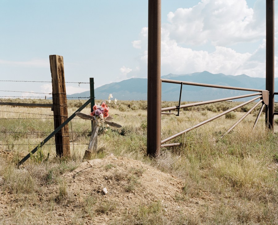 Flowers placed at a grave marker beside a ranch fence in New Mexico landscape with mountains.