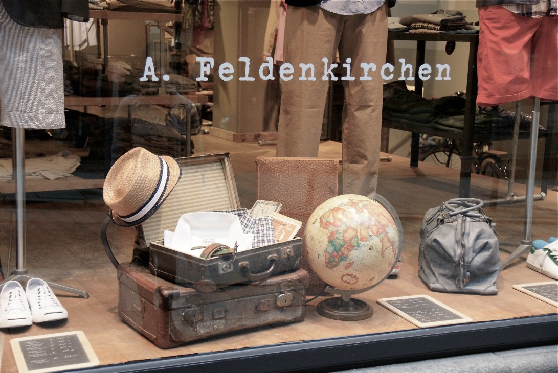 A. Feldenkirchen store window display featuring vintage suitcases, a straw hat, globe, and casual clothing on mannequins.