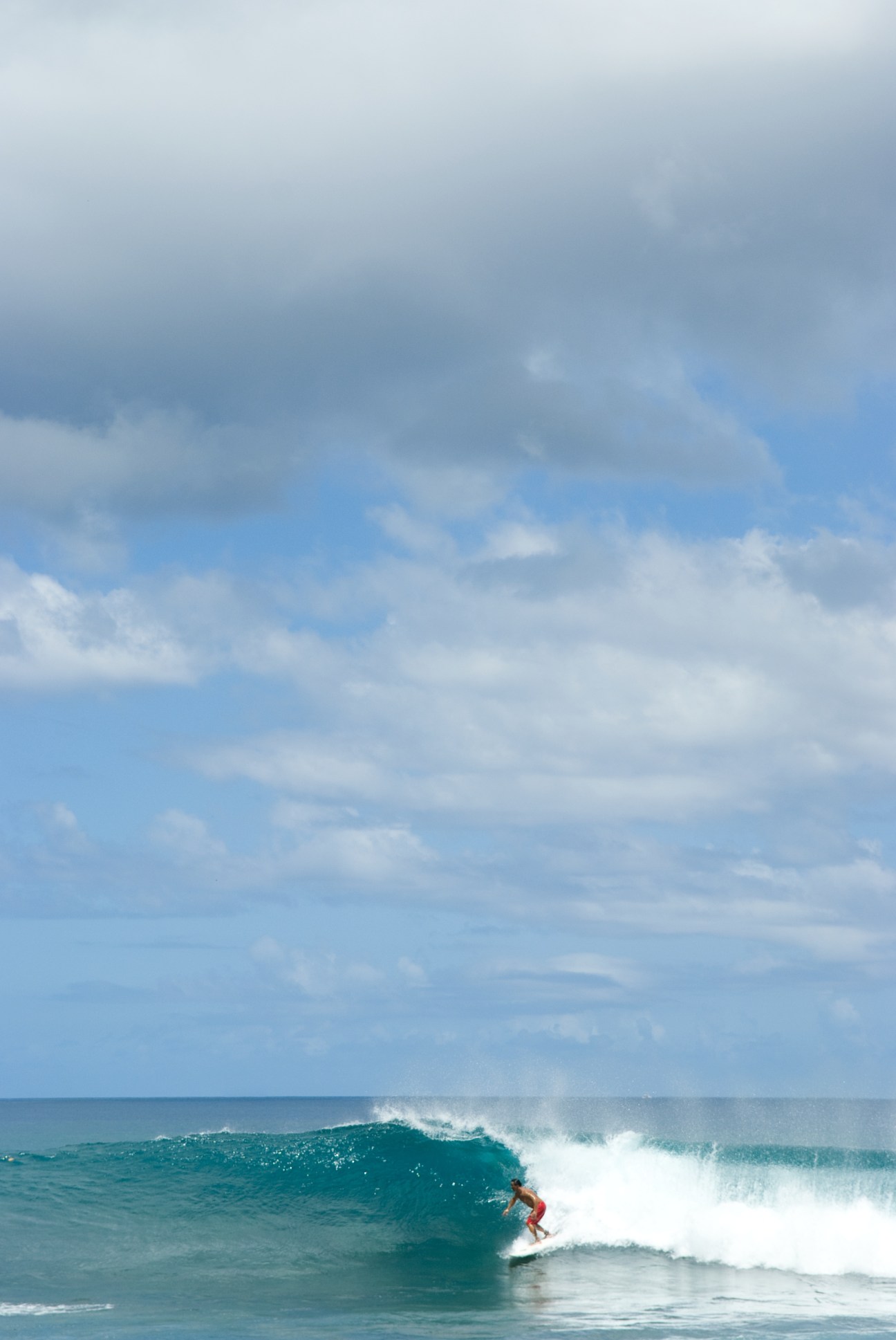 A surfer in red shorts rides a turquoise wave off Guam's coast under a partly cloudy sky.