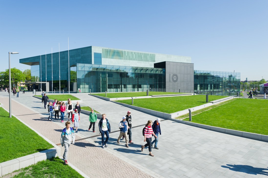 Modern glass and teal-colored museum building with green lawn and visitors walking on plaza.