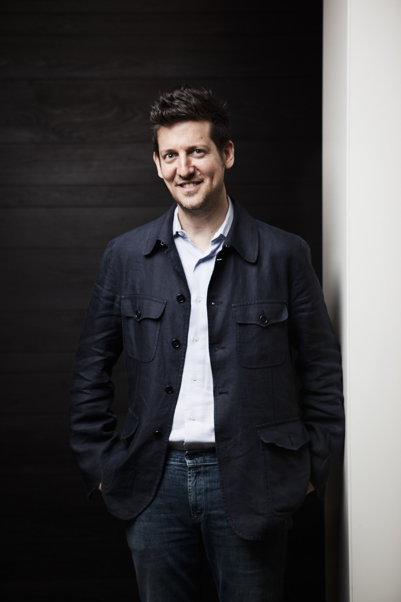 William Paley, head designer for Tony Chi, stands against a dark wall wearing a dark jacket and light blue shirt.