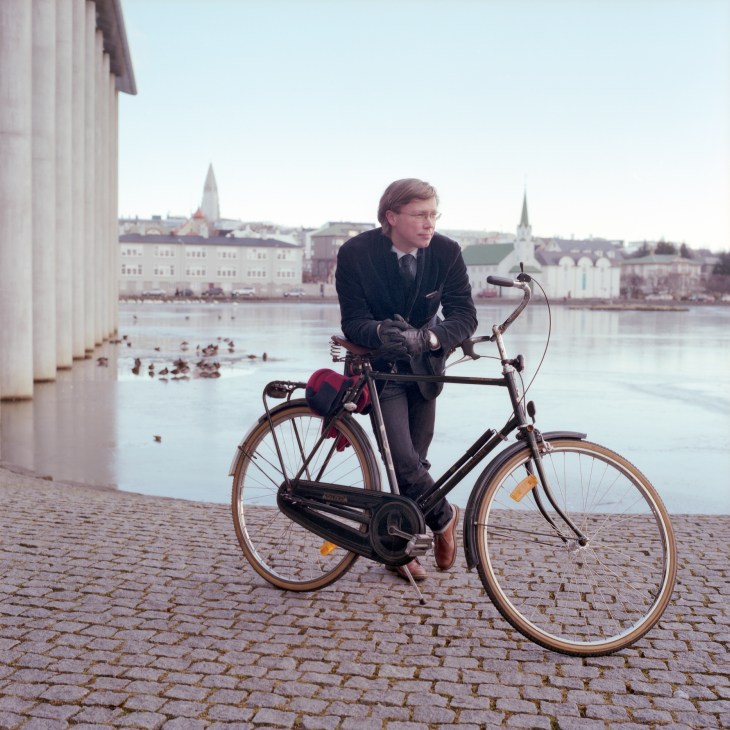City councillor Gísli Marteinn poses with a bicycle by the waterfront, with Reykjavik's churches and buildings visible across the water.
