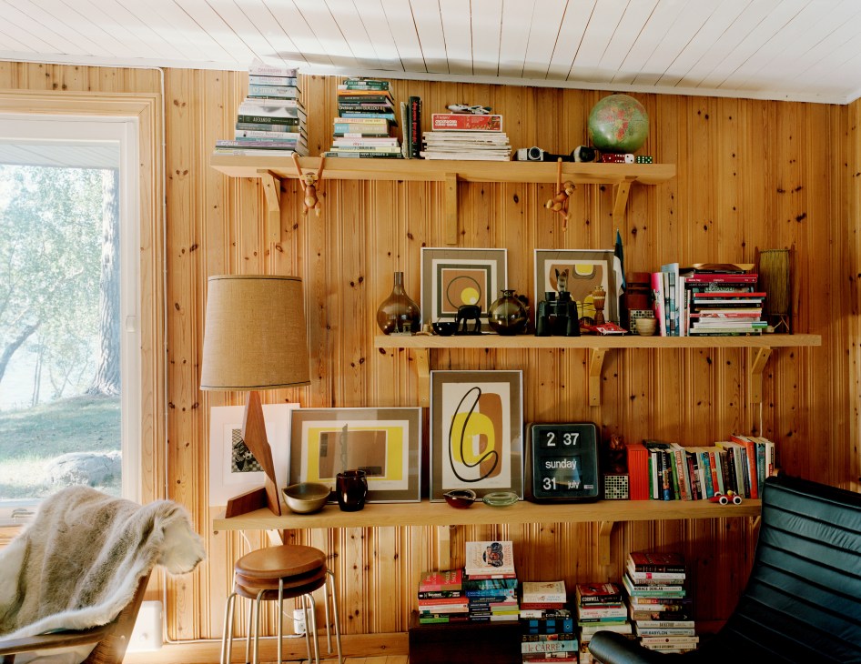 Wooden wall shelves display books, magazines, graphic prints, ceramic vessels, and decorative objects with a window view to the left.