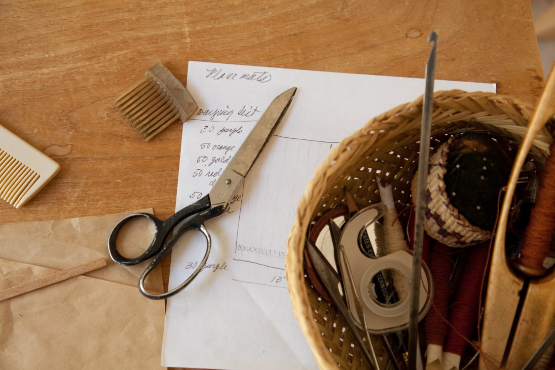 Weaving tools and supplies arranged on a wooden surface including scissors, combs, and a woven basket with thread.