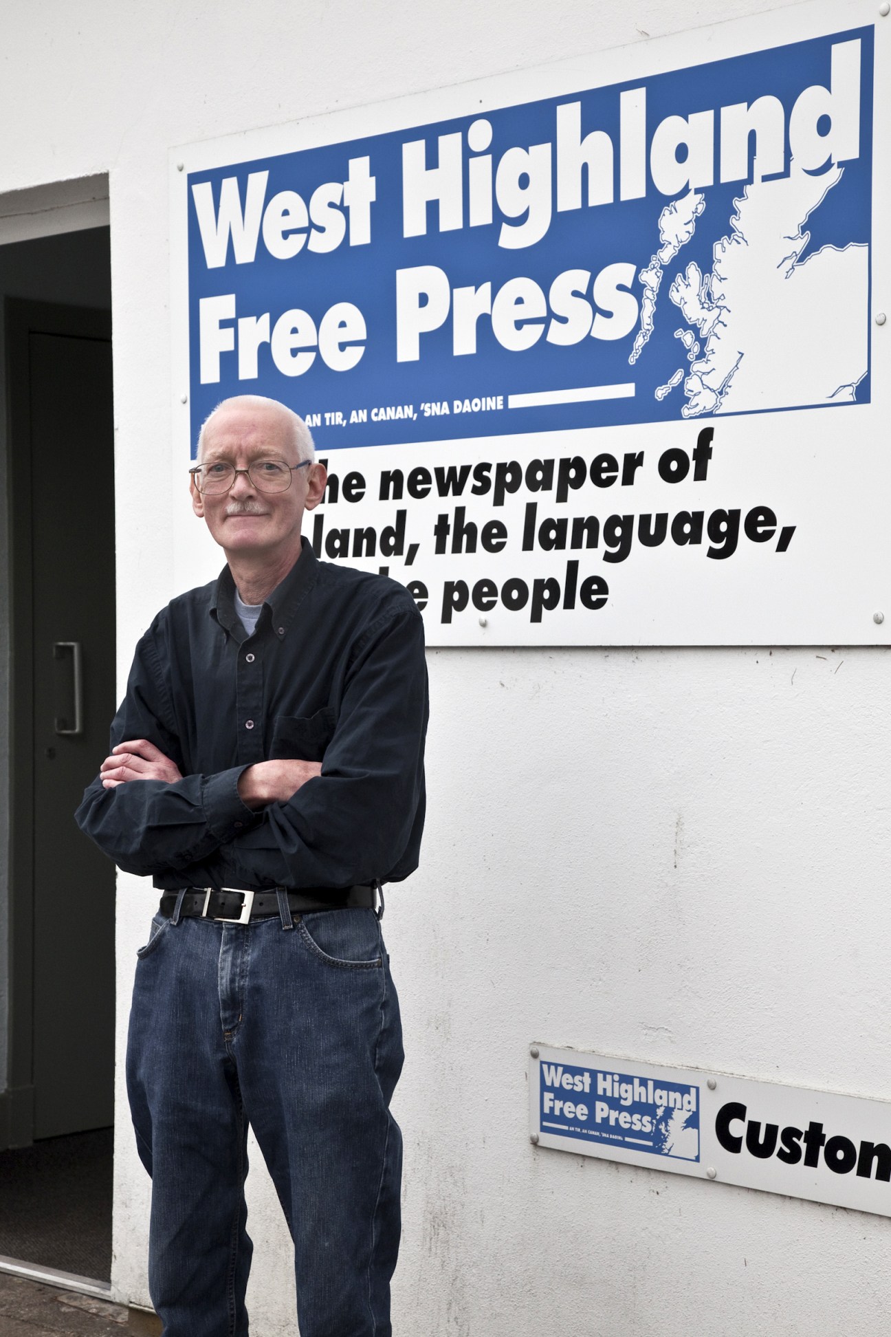 Editor Ian McCormack stands with arms crossed outside West Highland Free Press office in Scotland.