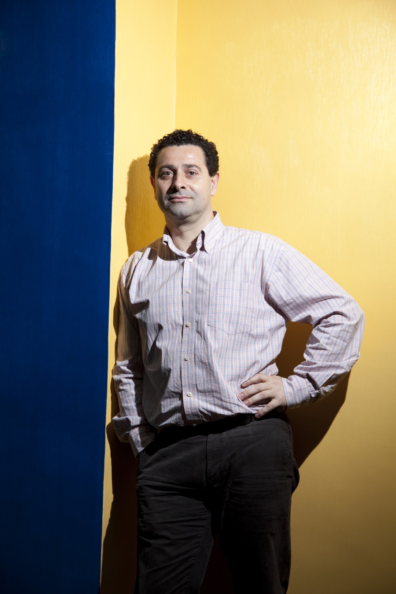 Portrait of Filipe Bragança, a car designer, standing against a blue and yellow wall wearing a striped shirt.