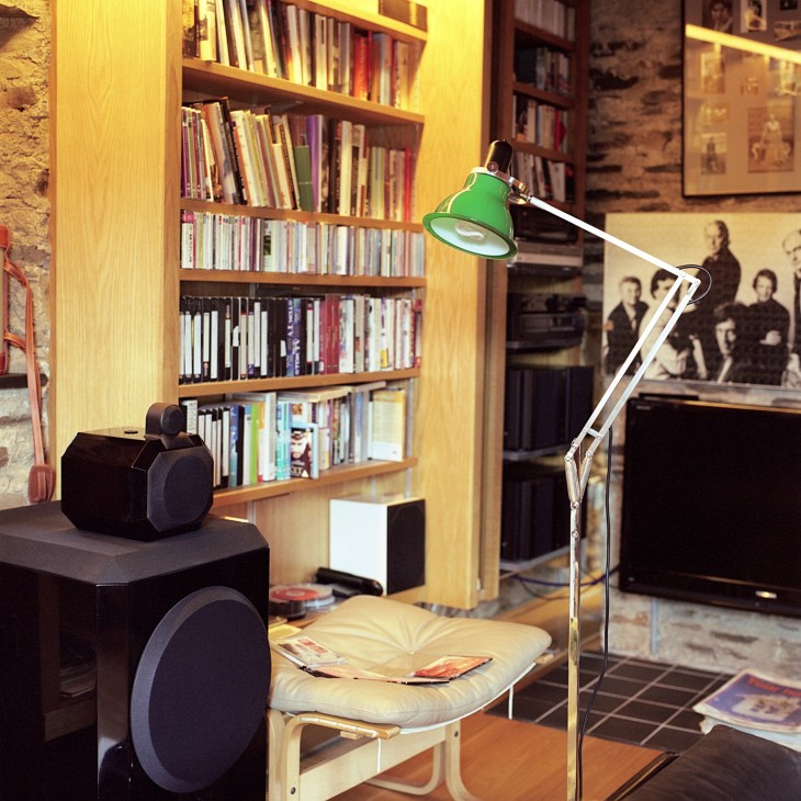 Green Anglepoise lamp beside B&W speaker in a room with wooden bookshelves and yellow walls.