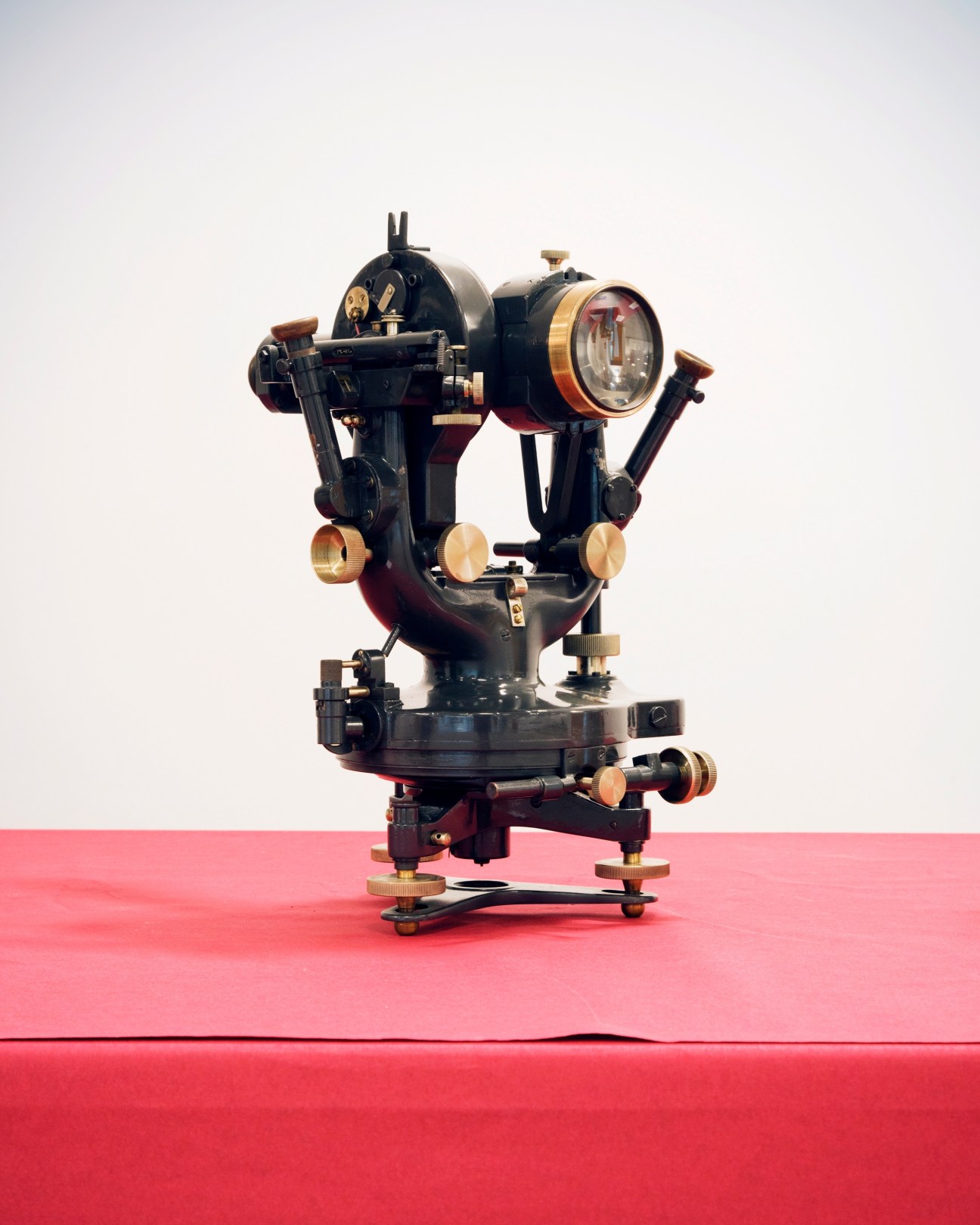 A vintage black and brass theodolite surveying instrument with compass and telescope on pink background
