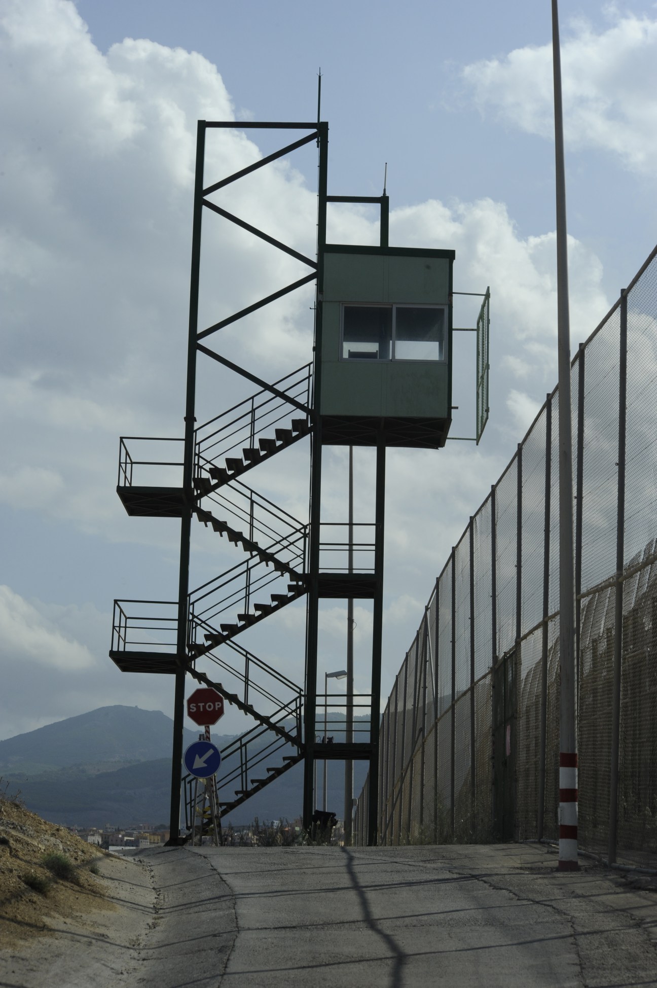 A tall metal sentry tower with stairs and observation booth beside a high metal border fence.