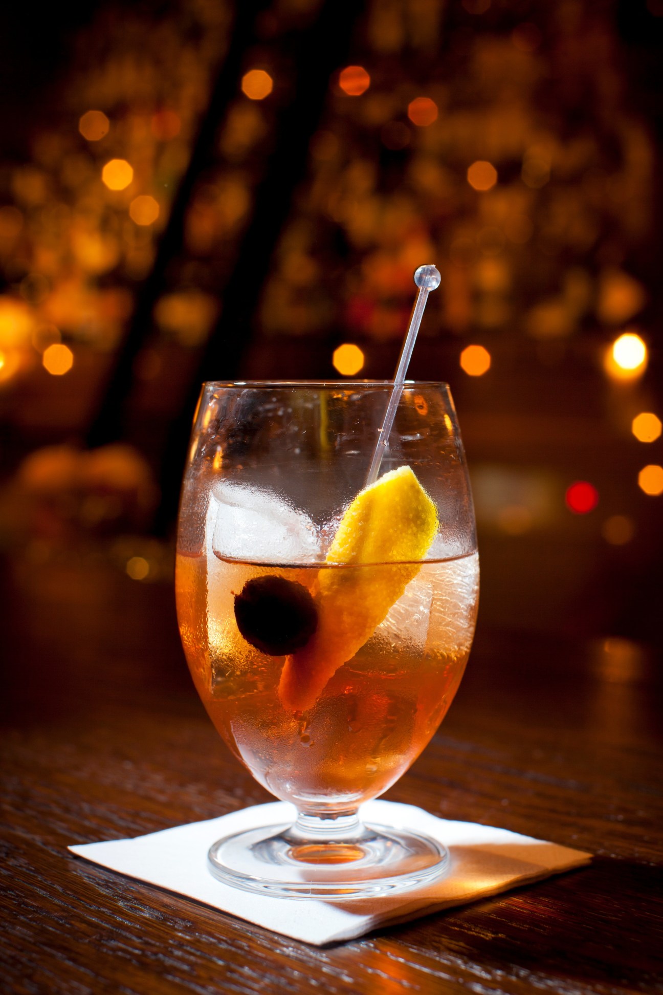 Amber cocktail with ice and lemon twist in a glass against blurred warm lights.