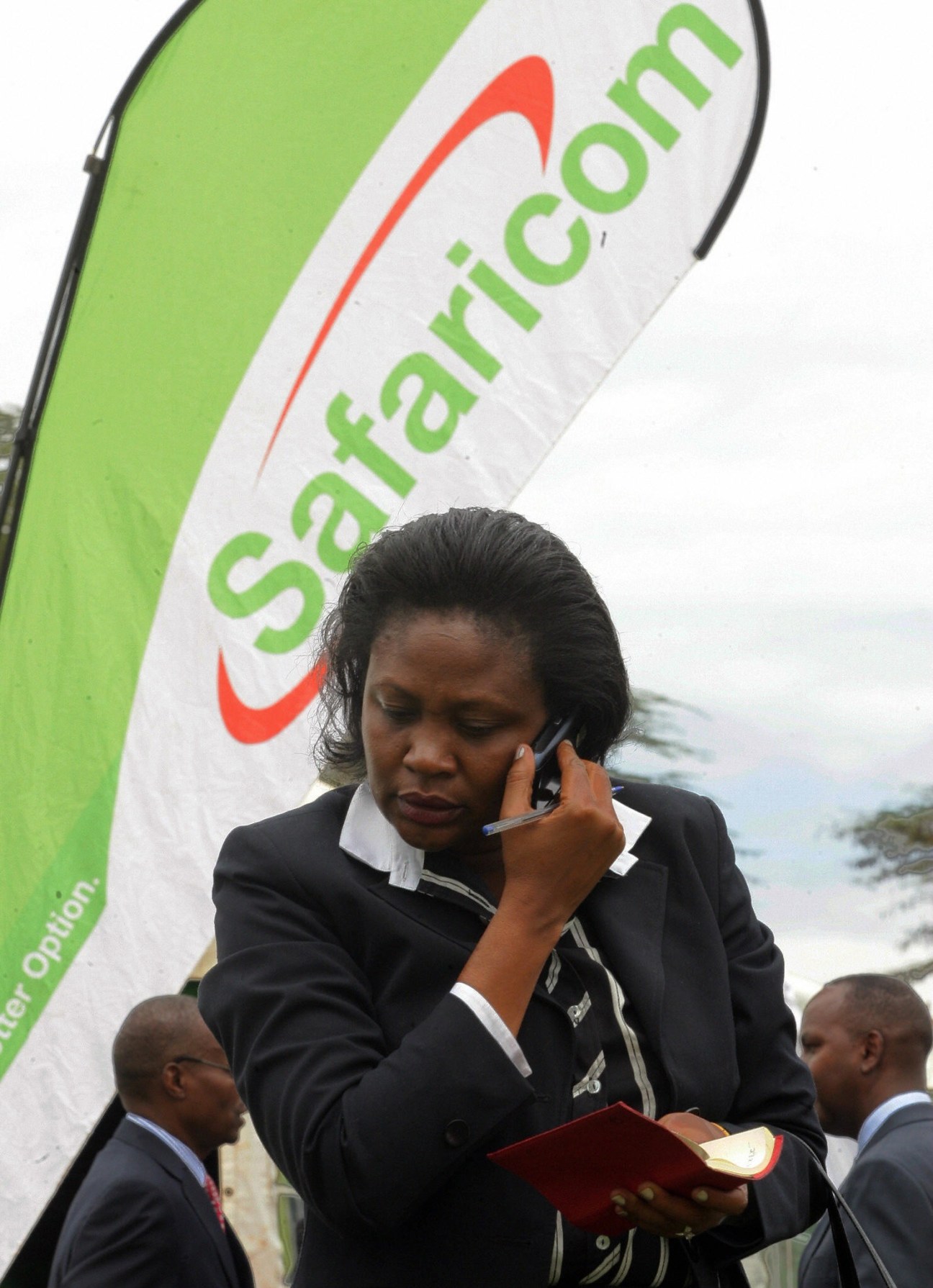 Woman in business attire using mobile phone in front of Safaricom banner.