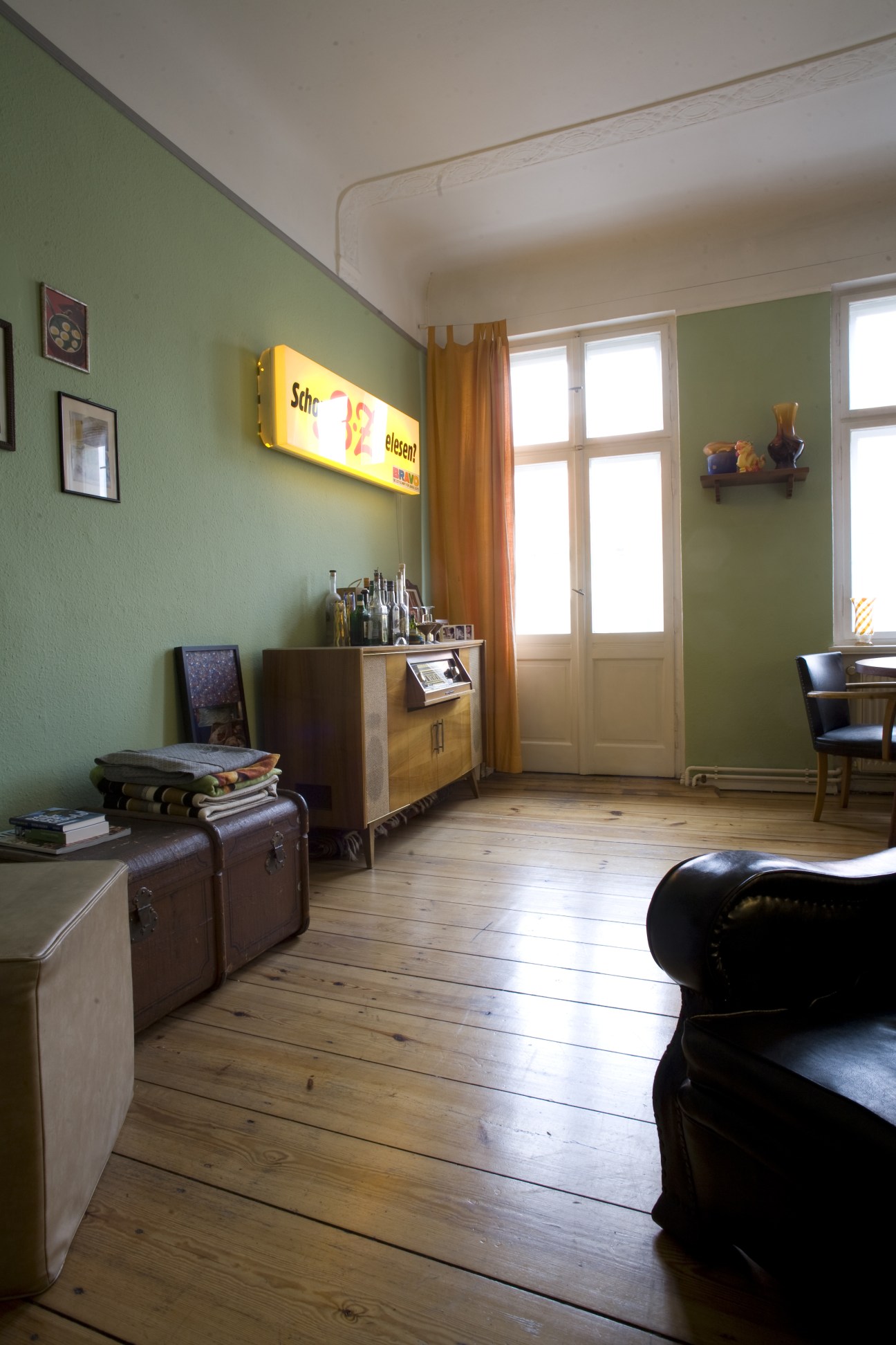 A living room with sage green walls, wooden floorboards, vintage furniture, and large windows with golden curtains.