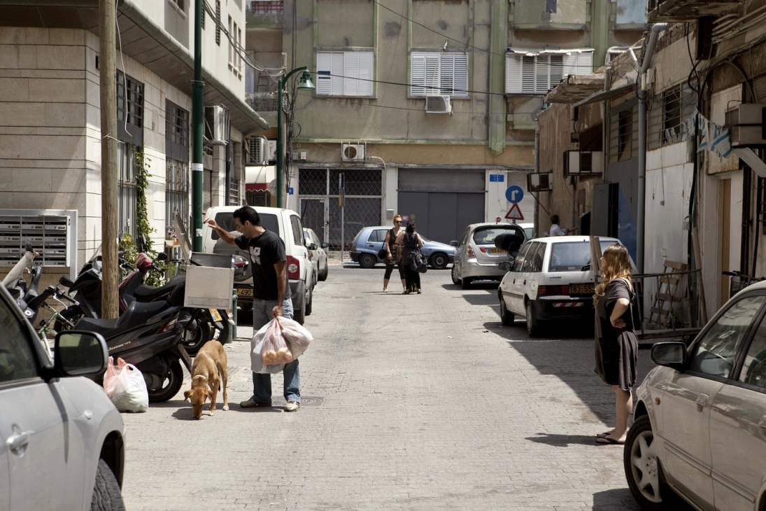A sunny Noga street with parked cars, pedestrians, and residential buildings with Middle Eastern architecture.