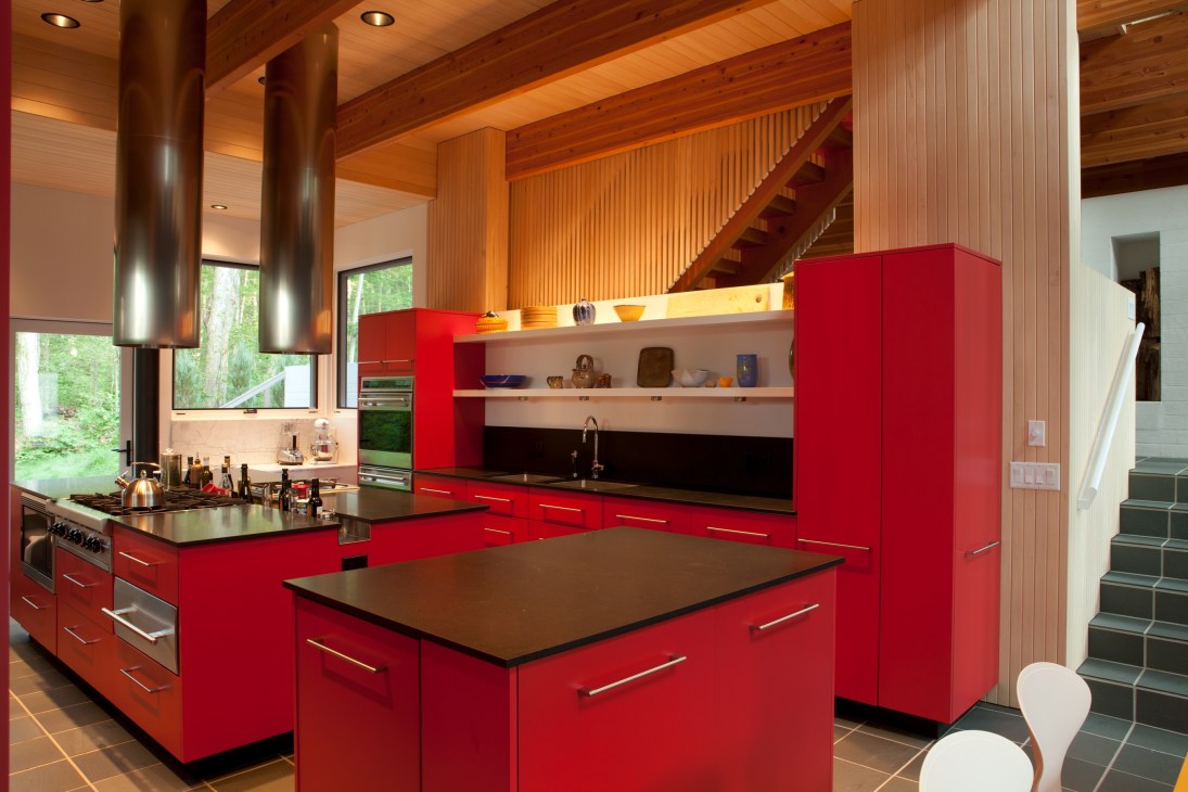 Modern open-plan kitchen with bright red cabinetry, black countertops, stainless steel appliances, and wooden beams overhead.