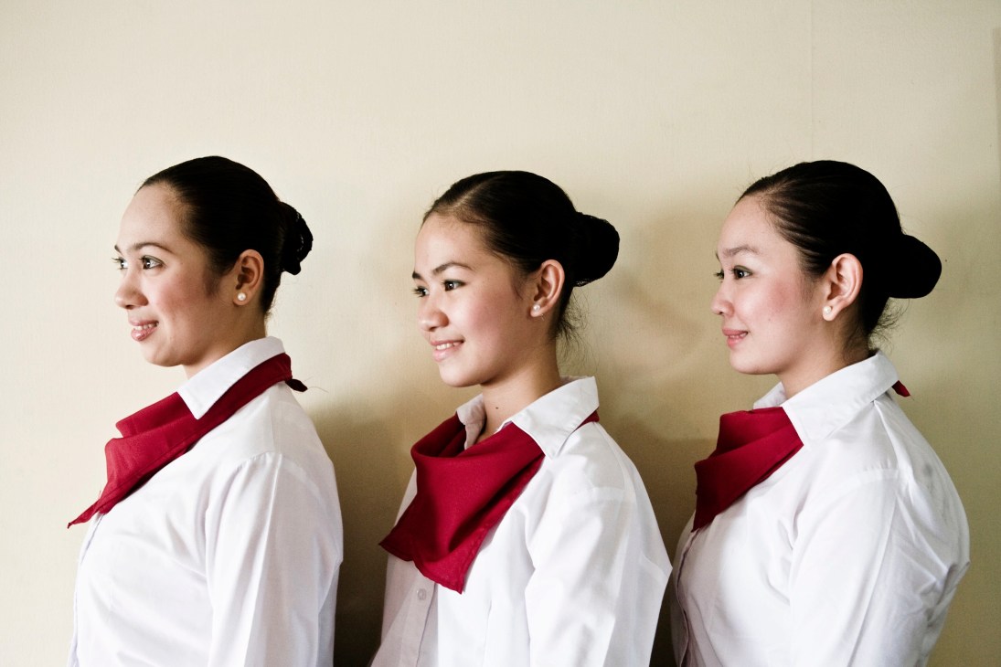 Three young Filipino women in white shirts and red scarves pose in profile against a beige wall.